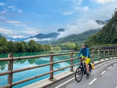 Radfahrer in blauer Jacke und gelber Hose auf Brücke über türkisfarbener Soča mit nebligen Bergen im Hintergrund, Slowenien.