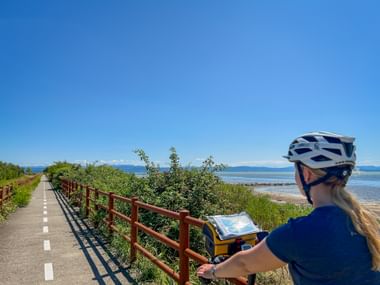 Radfahrerin mit Helm betrachtet Karte auf asphaltiertem Radweg mit Holzgeländer entlang der Adriaküste. Klarer blauer Himmel und ruhige See im Hintergrund.