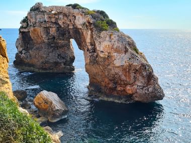 Natürliches Felsentor Es Pontàs im blauen Mittelmeer vor Mallorcas Küste, mit Vegetation oben und sichtbarer felsiger Küstenlinie.