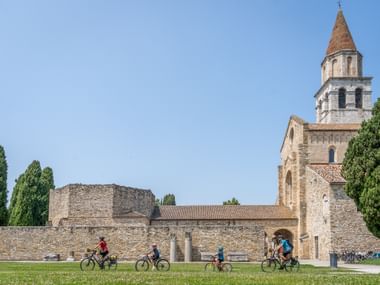 Family of four cycling on grass path past ancient stone walls and Santa Maria Assunta basilica with bell tower in Aquileia under blue sky.