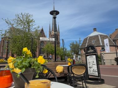 Außenterrasse eines Cafés mit gelben Blumen und Holzbänken in Woerden, mit historischem Kirchturm und traditionellen niederländischen Gebäuden.