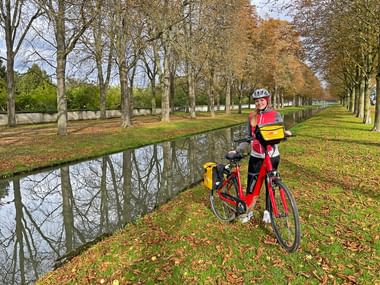 Radfahrer mit rotem Fahrrad und gelben Taschen steht neben einem Kanal mit Baumallee im Herbst. Herbstlaub bedeckt den Boden in der Pfalz.