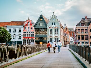 Fußgänger auf Brücke zu bunten historischen Giebelhäusern am Haverwerf-Ufer in Mechelen, Flandern. Kirchturm im Hintergrund sichtbar.
