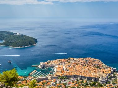 Aerial view of Dubrovnik's walled Old Town with terracotta roofs on a peninsula, marina, and forested island in the blue Adriatic Sea.