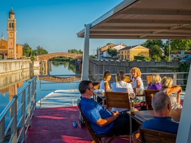 Passengers relaxing on the sun deck of MS Ave Maria in Portegrandi, with a church tower and bridge visible along the canal.