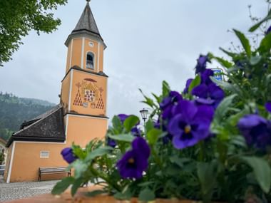 Orange St. Sebastian Church tower in Garmisch with pointed spire, framed by purple flowers in foreground and forested mountains in background.