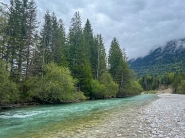 Turquoise Isar River flowing through pebble banks near Mittenwald, surrounded by coniferous forest with misty Alpine mountains in background.