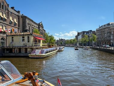 Amsterdamer Gracht mit Ausflugsbooten vor historischen Gebäuden. Traditionelle niederländische Architektur säumt beide Seiten unter blauem Himmel.