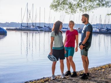 Drei Radfahrer mit Helmen machen Pause am Hafen des Starnberger Sees. Segelboote liegen im ruhigen Wasser, ferne Hügel unter klarem Himmel.
