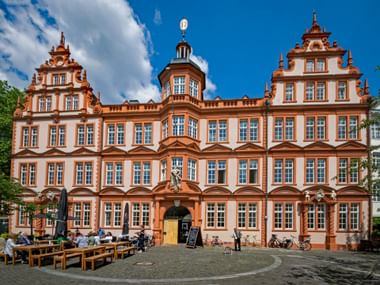 Orange-weißes barockes Gutenberg-Museum in Mainz mit verzierten Giebeln und Turm. Menschen sitzen an Tischen im Innenhof.