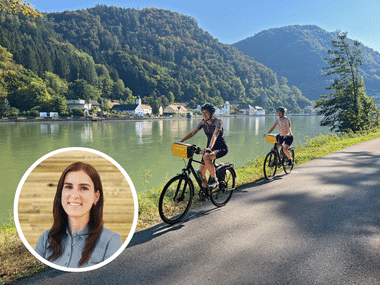 Two cyclists with yellow panniers riding along the Danube River between Linz and Krems, with forested mountains and a riverside village visible.