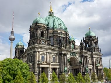 Berlin Cathedral with green copper domes and golden cross against cloudy sky. The TV Tower is visible on the left, with trees in foreground.