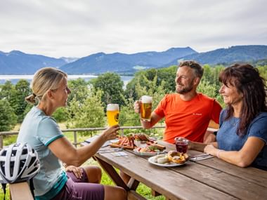 Three cyclists toasting with beer at a wooden table on a terrace. Lake Tegernsee and mountains visible in the background. Food platters on the table.