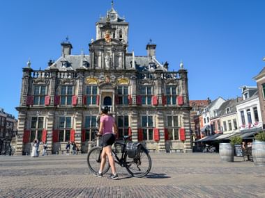 Radfahrerin mit Fahrrad vor dem prachtvollen Rathaus von Delft im Renaissance-Stil mit roten Bannern unter blauem Himmel.
