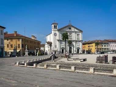 Piazza Grande in Palmanova with white cathedral facade, bell tower, and colorful buildings. The hexagonal square features stone patterns and benches.