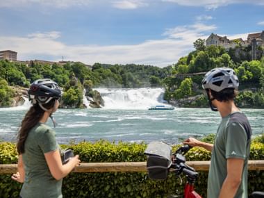 Zwei Radfahrer mit Helmen betrachten den Rheinfall bei Schaffhausen. Ein Ausflugsboot fährt im türkisfarbenen Wasser unterhalb des Wasserfalls.