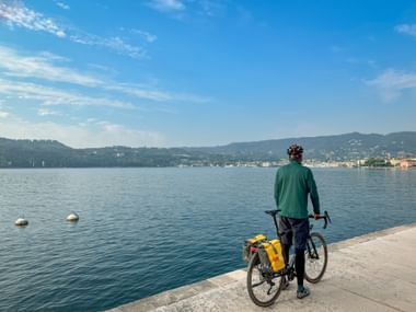 Cyclist with yellow panniers standing at waterfront promenade overlooking Lake Garda with hills and town of Salò in background under blue sky.