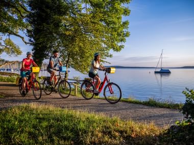 Three cyclists on a lakeside path at Starnberger See in Bavaria. Sailboats float on calm water, trees frame the scene under blue sky.