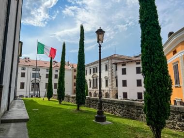 Innenhof in Udine mit grünem Rasen, hohen Zypressen, italienischer Flagge und historischen Gebäuden unter blauem Himmel mit weißen Wolken.