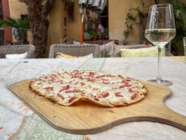 Flammkuchen mit Speck und Käse auf einem Holzbrett neben einem Glas Weißwein, serviert auf einer Außenterrasse in Weißenheim.