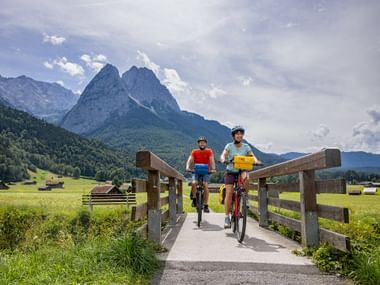 Two cyclists crossing a wooden bridge on a bike path in Bavaria near Garmisch, with dramatic mountain peaks and green meadows in the background.