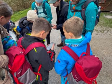 Group of hikers in Köstendorf gathered around a smartphone, with children wearing red backpacks and adults in turquoise jackets.