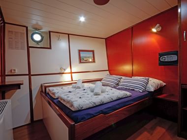 Doppelkabine auf der MS Macek mit Holzbett, gestreiften Kissen, Bullauge und warmer Holzvertäfelung. Ein Safe ist an der roten Wand montiert.