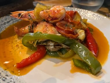 Traditioneller portugiesischer Cataplana-Fischeintopf mit Garnelen, Muscheln, Paprika und Tomaten in orangener Brühe auf weißem Teller.