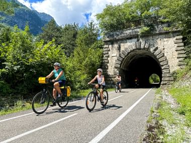 Drei Radfahrer nähern sich einem Steintunnel auf der Alpe-Adria-Radroute bei Dogna, umgeben von grünen Bäumen und Bergen.