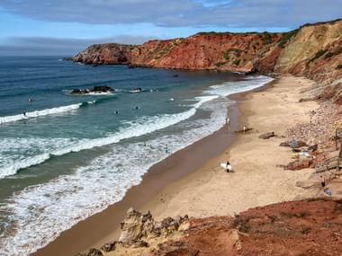 Sandstrand Praia do Amado mit Surfern in den Wellen, eingerahmt von dramatischen rot-gelben Klippen an der portugiesischen Atlantikküste.