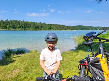 Smiling child with helmet sitting by bicycle on grassy lakeshore at Starnberger See. Blue lake and forest in background under clear sky.