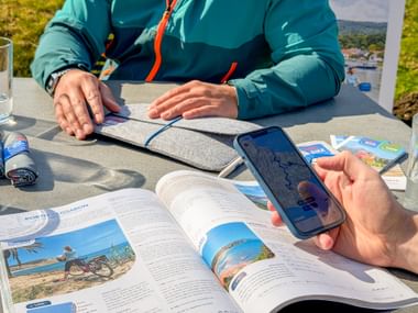 Zwei Personen an Tisch im Freien betrachten Radreisekatalog und Smartphone mit Routenkarte. Reisebroschüren und Wasserflasche auf dem Tisch.
