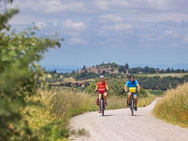 Two cyclists riding on a gravel path through golden fields in Val d'Orcia, Tuscany. A hilltop village with cypress trees is visible in the background.
