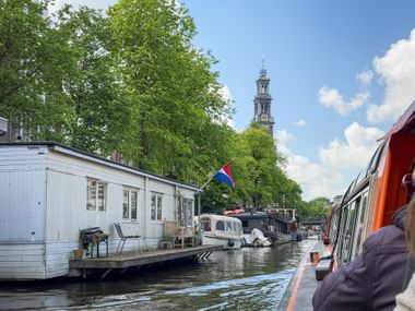 Blick von einem Kanalboot auf der Prinsengracht in Amsterdam mit Hausbooten, grünen Bäumen und dem Westerkerk-Kirchturm unter blauem Himmel.