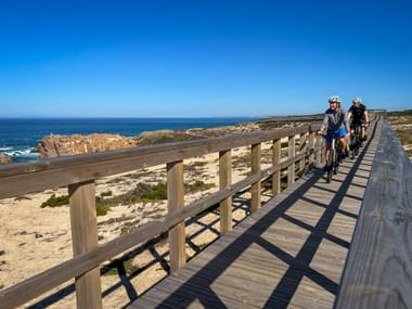 Two cyclists riding on a wooden boardwalk along the coast in North Portugal. The path has railings and runs beside sandy beach and blue ocean.