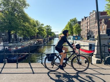 Radfahrerin auf Brücke über die Brouwersgracht in Amsterdam mit historischen Giebelhäusern, Hausbooten und Bäumen entlang des Kanals.