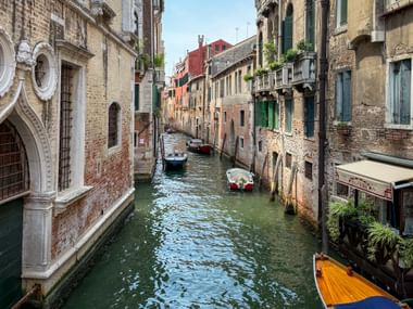 Schmaler venezianischer Kanal mit vertäuten Booten zwischen historischen Gebäuden. Verwitterte Fassaden in Beige- und Rottönen säumen den Wasserweg.