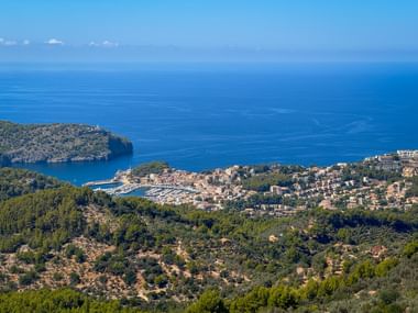 Luftaufnahme der Bucht von Port de Sóller auf Mallorca mit Hafen, rotgedeckten Gebäuden, bewaldeten Hügeln und tiefblauem Mittelmeer.