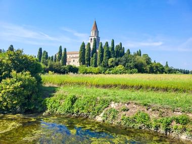 Historische Kirche in Aquileia mit hohem Glockenturm, umgeben von Zypressen, über grüne Felder und einen Bach hinweg unter blauem Himmel.