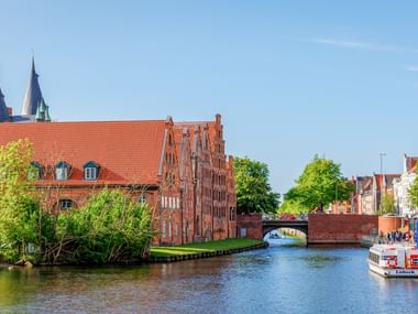 Rote Backsteingebäude der Salzspeicher am Kanal in Lübeck mit Kirchtürmen, Brücke und Ausflugsboot unter blauem Himmel.