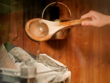 Hand pouring water from a wooden ladle onto sauna stones, with a copper bucket in the background. Water drips from the ladle onto white stones.