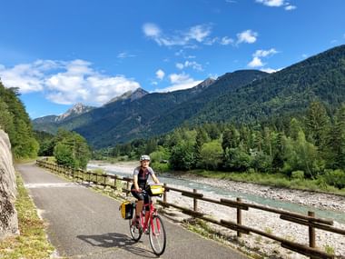 Radfahrer mit rotem Fahrrad und Packtaschen auf asphaltiertem Weg neben Fluss, mit Holzzaun, bewaldeten Bergen und blauem Himmel in Kärnten.