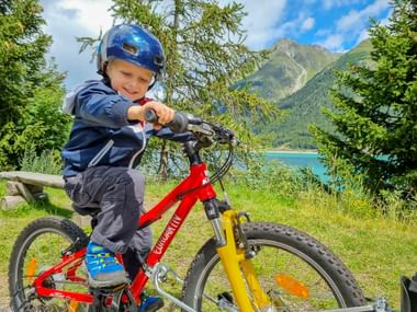 Kleines Kind auf rotem Fahrrad mit blauem Helm am Reschensee. Türkisfarbener See und grüne Berge im Hintergrund unter blauem Himmel.