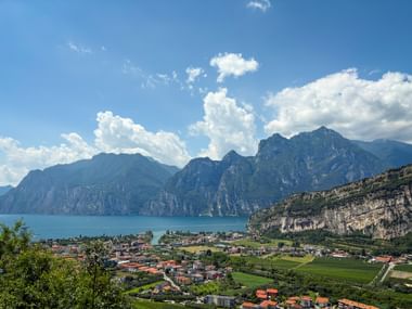 Panoramic view of Lake Garda with steep mountains, a lakeside town with red roofs, green fields, and dramatic cliffs under a blue sky with clouds.