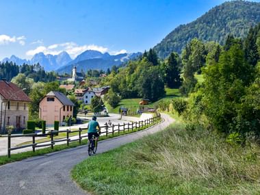 Radfahrer in türkisfarbener Jacke auf asphaltiertem Weg bei Coccau mit Alpendorf, Kirchturm und Berggipfeln im Hintergrund.