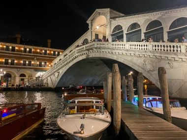 Beleuchtete Rialtobrücke in Venedig bei Nacht mit Booten am Holzsteg im Canal Grande. Menschen sind auf der Balustrade der Brücke sichtbar.