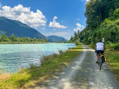 Cyclist on gravel path along turquoise Drau River with mountains and forest. Blue sky with white clouds above the Alpine landscape.