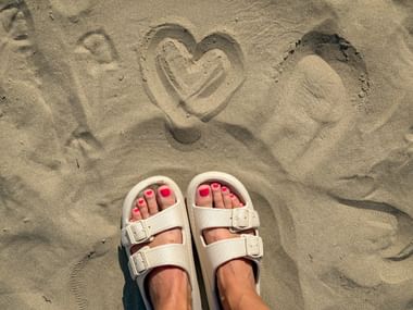 Füße in weißen Sandalen mit rosa Nagellack am Sandstrand in Caorle. Ein Herz ist in den Sand über den Füßen gezeichnet.
