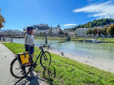 Cyclist with helmet standing beside bike on grassy riverbank of Salzach River in Salzburg, with historic old town and fortress visible across the water.