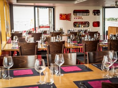 Bright restaurant interior on MS Ave Maria with wooden tables set with wine glasses and pink napkins. Large windows show river views.